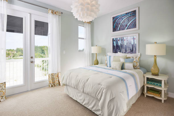 A large bed sits next to a double door leading out to a small balcony overlooking a sandy seascape. The wall is a blue gray color, and the doors and windows are white. The curtains are see through with fringe orange and blue patterning. On the ceiling is an interesting chandelier of white overlapping cups, creating a flower effect. On the wall above the bed hangs two photos of back, black bicycle tires, blue tire guards, and red, oval reflectors. Below, on the bed, sits two long white pillows, two pillows matching the drapery, and three pillows matching the white, light blue, and tan bed spread. On the four leaf clover, sandy white end tables sits a yellow lamp with a twist in its neck and a white lamp shade. Three books of various sizes and colors are on the end table in shot. The floor is a brown carpet.